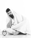 Man with calm, bored face sits under blanket near alarm clock. Man wants to stay in bed, white background.