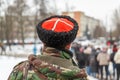 man in camouflage jacket and cossack hat with white cross on red watching blurry crowd of people