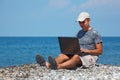 Man in cap sitting on beach with laptop on knees Royalty Free Stock Photo