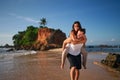 Couple enjoys piggyback on sandy beach. Man carries girlfriend on back, smiles by sea near cliff. Holidaymakers share