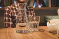 Man in checkered shirt sitting at wooden table with sugar bowl, glass and jud with water in cafe