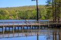 A man and a child riding bikes across a long brown wooden bridge over a rippling blue lake surrounded by lush green trees