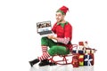 man in christmas elf costume sitting on sleigh and holding laptop with depositphotos website on screen isolated