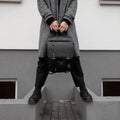 Man in a classic fashionable plaid coat in stylish boots in black trousers with a trendy leather black backpack stands near a wall Royalty Free Stock Photo