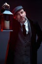 A man in a classic suit, coat and cap. Stands. Holds a vintage lantern in his hand.