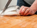 Man cleaning fresh sea bream on a wooden board with knife holding fish by it head. Descaling stage