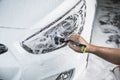A man cleans the left headlight cover of a white sedan covered in car shampoo with a fine brush. A vehicle being serviced at a