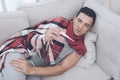 A man with a cold sits on the couch, hiding behind a red rug. He measures the temperature with a digital thermometer Royalty Free Stock Photo