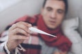 A man with a cold sits on the couch, hiding behind a red rug. He measures the temperature with a digital thermometer Royalty Free Stock Photo