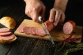 A man cuts veal sausage on a wooden cutting board. Preparing delicious sandwiches for dinner on the kitchen table at home Royalty Free Stock Photo