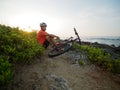 Man cyclist wearing white helmet and orange t-shirt seating with a mountain bike, at ocean shoreline. Royalty Free Stock Photo