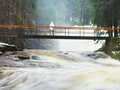 Man with dog on bridge over troubled water. Huge stream of rushing water masses below small footbridge. Fear of floods.