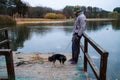 Man dog owner and his friend dog are standing on the wooden pier near lake and enjoying the landscape during their walking Royalty Free Stock Photo