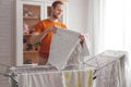 Man doing home chores. Caucasian man removes clothing and baby sheets after laundry from portable dryer in living room