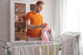 Man doing home chores. Caucasian man removes clothing and baby sheets after laundry from portable dryer in living room