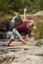 Man doing the triangle pose on rocks by a waterfall