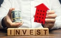 A Man With Dollars And A House Figurine And The Word Invest. Real Estate Financial Investment. Favorable Investment Climate, Cheap