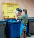 Man dressed in green shirt and blue apron is throwing away paper box with garbage into trash.