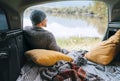 A man dressed in warm knitted clothes sitting in the cozy camper trunk and enjoying the mountain lake view. Warm early autumn auto