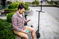 Man with e-scooter relaxing and using smartphone on bench in city. Modern male with electro scooter sitting on bench in park while Royalty Free Stock Photo