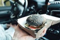 Man is eating black burger with meat, fish, tomato, salad, sause in car. Fast, safe, takeaway food on the road to coronavirus Royalty Free Stock Photo