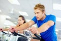 Man on exercise bike. Young man cycling at the gym with woman in background.