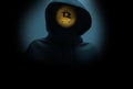 Man without a face in a hood holds a luminous bitcoin icon on a dark background.