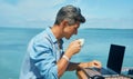 Man freelancer working at morning on the beach by the sea, drinking coffee, using laptop computer. Royalty Free Stock Photo