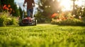 A man, gardener mowing the lawn with red lawn mower, green grass and vegetation, flowers on it with blue sky in the Royalty Free Stock Photo