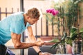 Man gardening touching the leaves of a plant on a table. In the vase there is a purple orchid. The young man is wearing a light