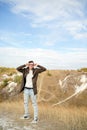 Man with glasses in jeans, t-shirt and cape stands in hilly terrain. Adult male in hood adjusts his eyeglasses and