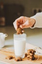 A man is going to dip a oatmeal cookie in the form of a gingerbread man into a glass. There is a glass of milk on the kitchen