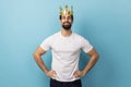 Man in gold crown, standing with hands on hips, looking at camera with conceited expression.