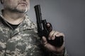 A man with a gray beard and in camouflage blows on the barrel of a pistol. Military conflicts, murders and crimes. Grey background Royalty Free Stock Photo