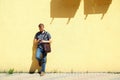 Man in gray polo shirt and jeans with leather bag standing near bright yellow wall and using smartphone. Full length portrait of