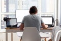 A man in a gray t-shirt is sitting at a workplace with two laptops and a monitor near the window. Remote work from home