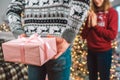 Man in grey Christmas sweater stands in front of woman and hide present form her. She looks excited. Young woman keeps