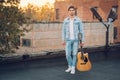 Young man holding guitar in city on sun rays background. Handsome hippie style guitarist engrossed on music outdoors. Concept of