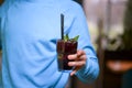 Man hand holding a drink in a tall glass. Refreshing cocktail with mint and plastic straw. Person in blue sweater.