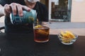 Man hand pour or fill drink in glass with ice and lemon, plate with chips over a black table in a bar or caf