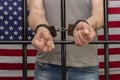 A man is handcuffed in a cell behind bars against the background of an American flag. Concept: the prisoner in the courtroom, the Royalty Free Stock Photo