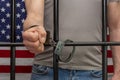 A man is handcuffed in a cell behind bars against the background of an American flag. Concept: the prisoner in the courtroom, the Royalty Free Stock Photo