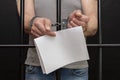 A man in handcuffs holds a white sheet of paper in his hands behind bars in a cell. Concept: a petition for clemency, the last wor