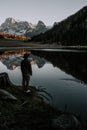 A man with a hat is standing by a mountain lake in the Dolomites. The mountains and the rest of the landscape are reflected in the Royalty Free Stock Photo