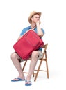 Man in a hat, t-shirt, shorts and slippers, sitting on a chair, waiting, hugging a suitcase. Isolated on white background