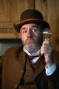 A man in a hat and a Victorian costume holds a incandescent lamp in one hand and looks at it with surprise.