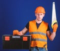 Man in helmet, hard hat carries toolbox and holds handsaw, blue background. Worker, repairer, repairman on serious face Royalty Free Stock Photo
