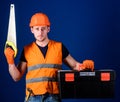 Man in helmet, hard hat carries toolbox and holds handsaw, blue background. Worker, repairer, repairman on serious face Royalty Free Stock Photo