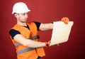 Man in helmet, hard hat holds building plan, controls works, red background. Engineer, architect, builder on strict face Royalty Free Stock Photo