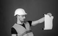 Man in helmet, hard hat holds building plan, controls works, red background. Engineer, architect, builder on strict face Royalty Free Stock Photo